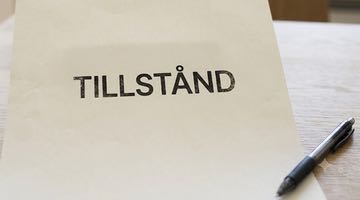 Ett vitt papper som ligger på ett träbord med ordet ”TILLSTÅND” tryckt i centrerad, svart versal text. Bredvid pappret ligger en svart penna.